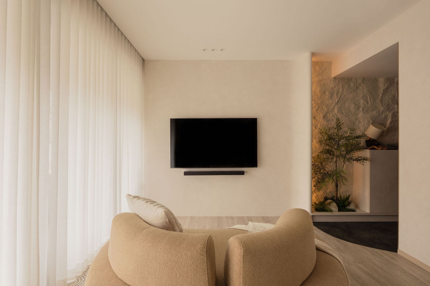 Modern living room with wall-mounted TV, soundbar, beige curved sofa, sheer curtains, and natural stone accent wall.