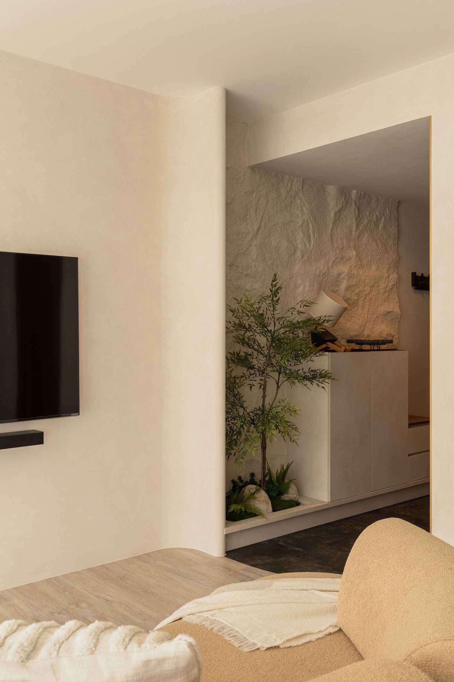 Modern living room with wall-mounted TV, beige sofa, and an open doorway revealing a plant and textured stone wall.