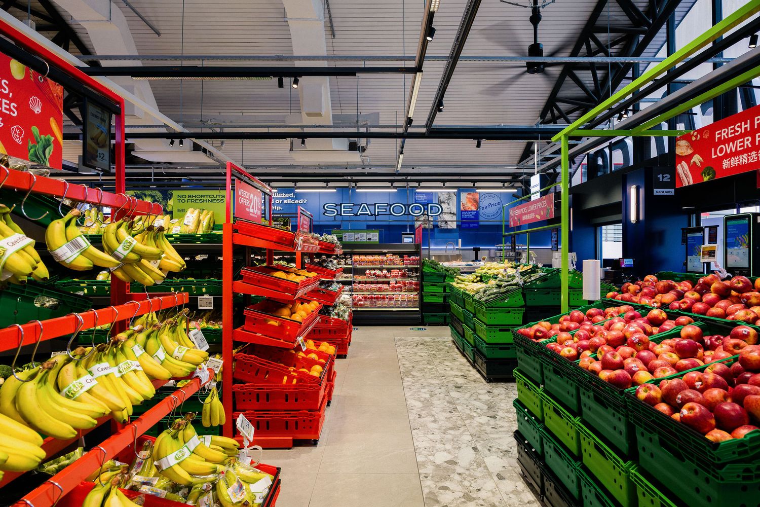 Fresh produce section in a grocery store with bananas on red shelves and crates of red apples on green display stands.