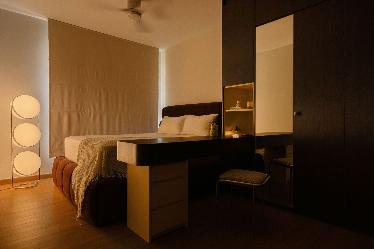 Cozy modern bedroom with warm ambient lighting, a bed with neutral linens, a floating wood vanity desk, a mirrored wardrobe and a stacked gl