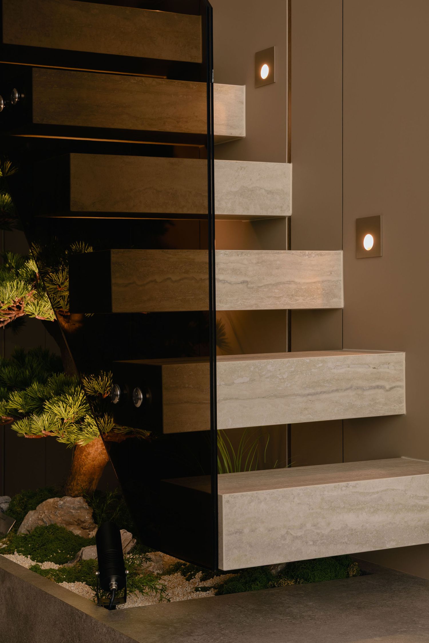 Floating travertine stone staircase with glass balustrade, recessed wall lights, and a pine bonsai garden with rocks and moss at dusk.