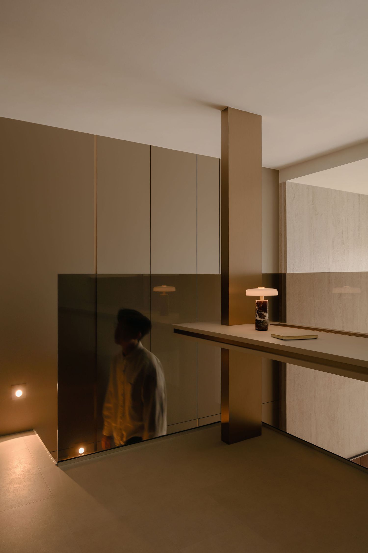 Modern minimalist interior with warm taupe panels, a glass balustrade reflecting a person, and a small table lamp on a floating shelf.