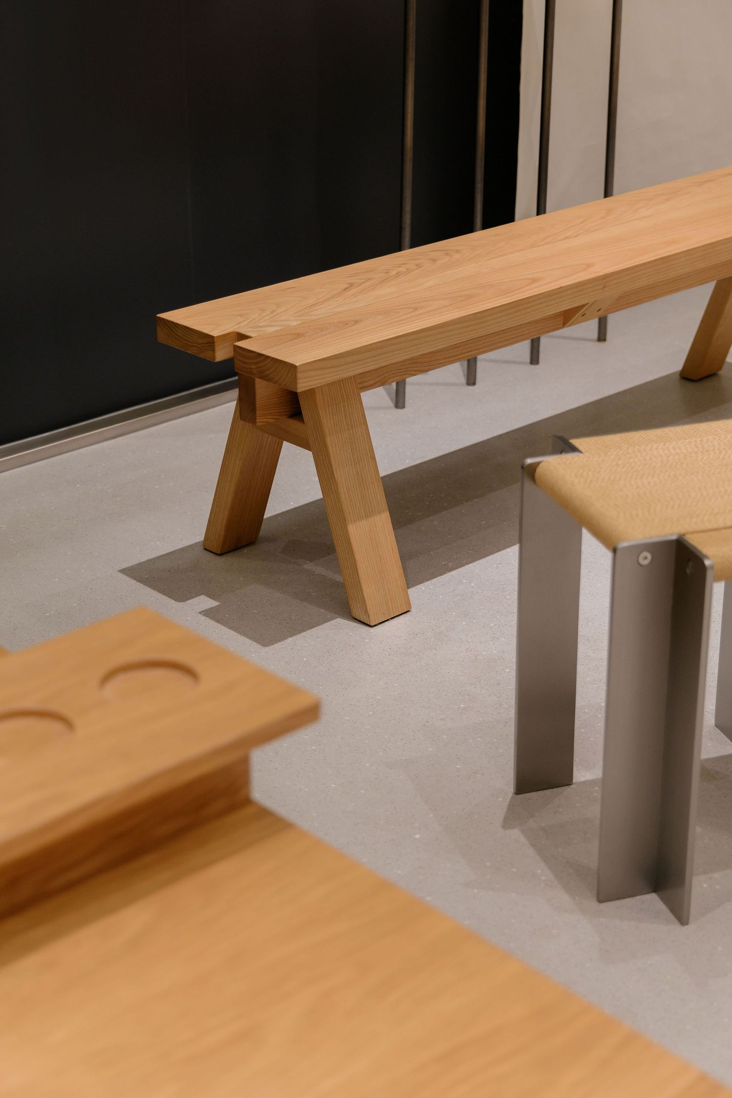 Minimalist oak wooden bench with splayed legs beside a modern stool with steel legs on a polished concrete floor.