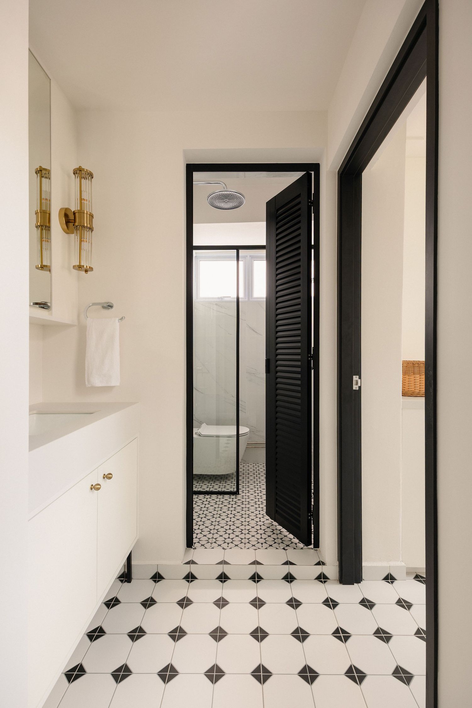 Black and white patterned tile bathroom with white vanity, brass fixtures, and black louvered door leading to shower.