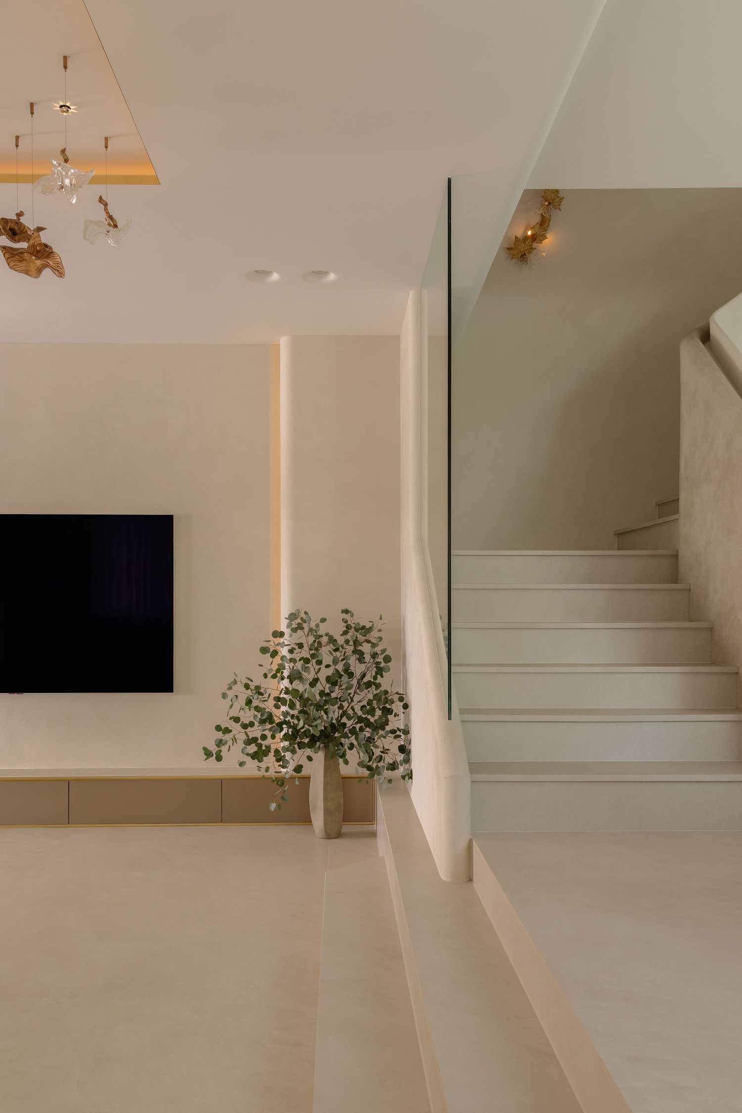 Modern minimalist living room with beige walls, built-in TV unit, green plant, and elegant staircase with glass railing.