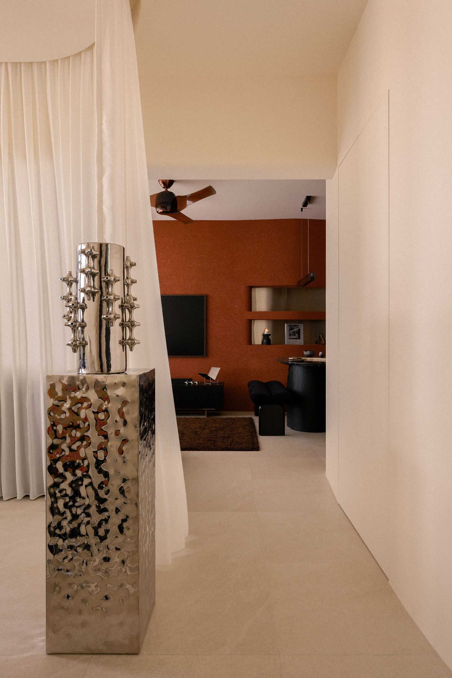 Modern hallway with a hammered silver metallic pedestal and sculpture, sheer white curtains, and a terracotta-walled living area beyond.