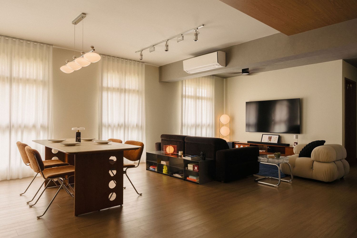 Open-plan living room with wooden dining table and chairs, dark sofa, wall-mounted TV, sheer curtains and warm hardwood flooring.