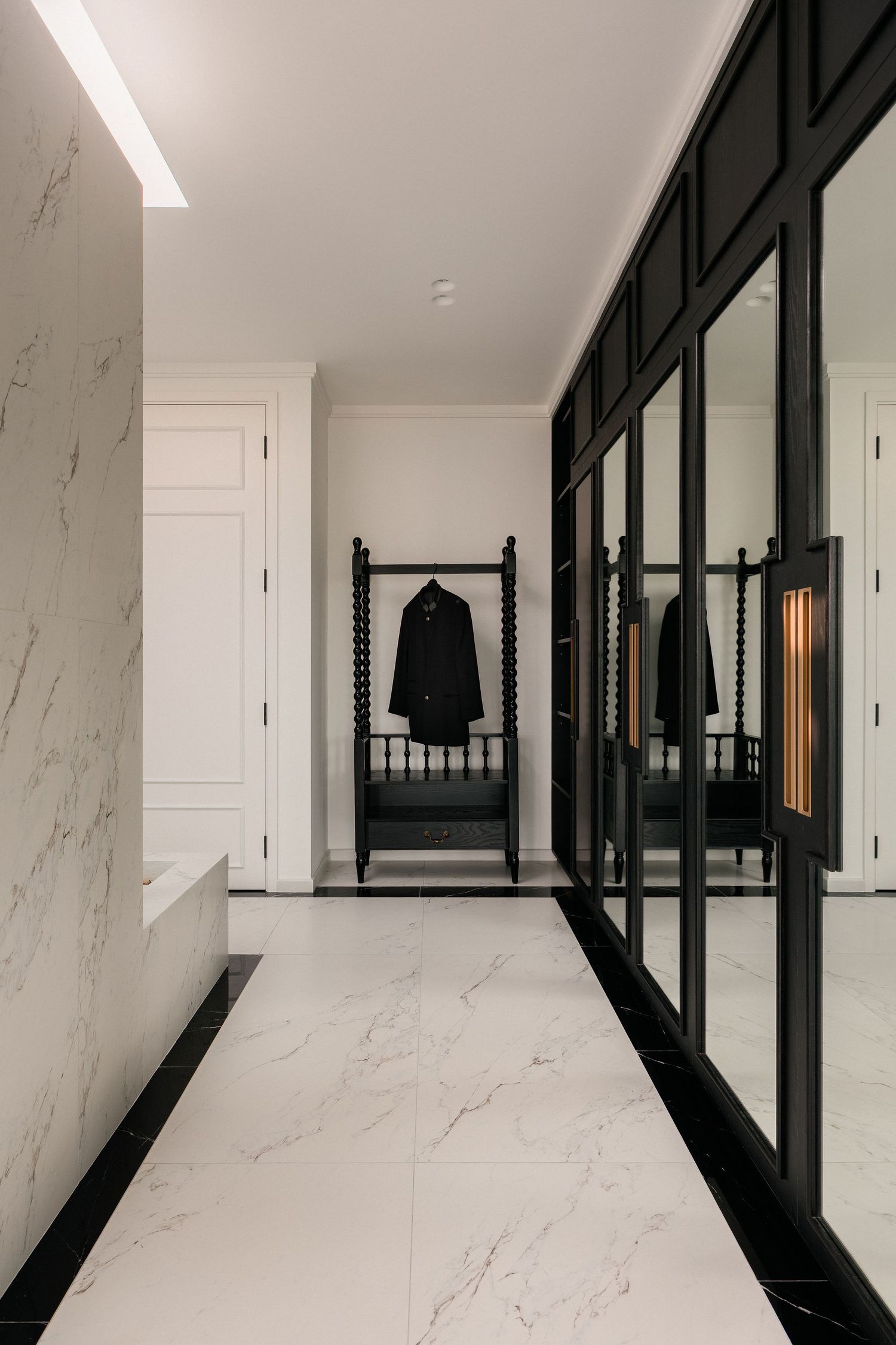 Elegant hallway with white marble floors and walls, black mirrored wardrobe doors, and a dark bench with a coat hanging on a stand.