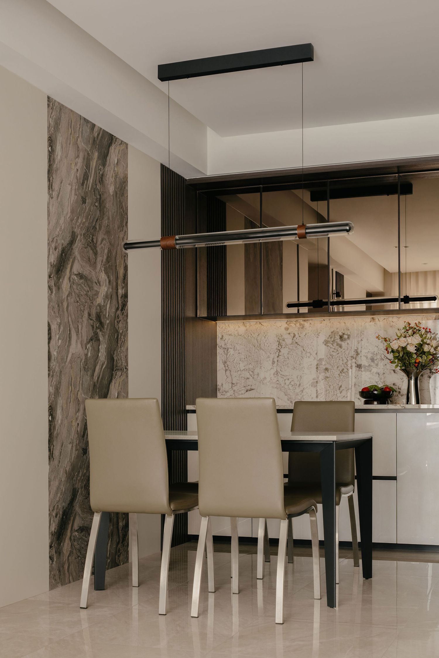 Modern dining area with four beige leather chairs, a dark table, linear pendant light, marble accent wall and mirrored backsplash shelf.