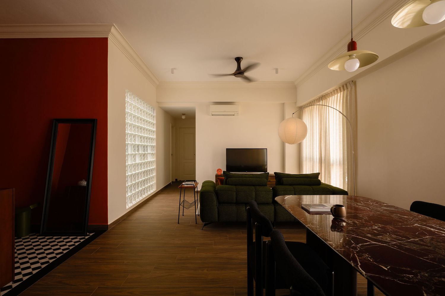 Modern open-plan living room with a red accent wall, glass block window, green sofa, ceiling fan and dark marble dining table.