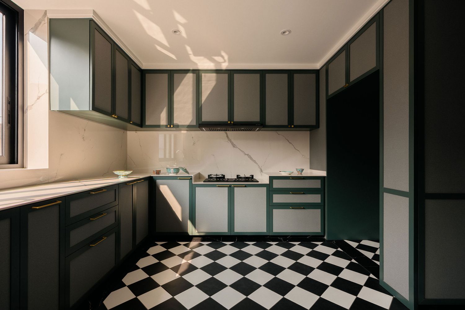 Modern kitchen with dark green cabinets, white marble backsplash and black and white checkered tile floor lit by warm sunlight.