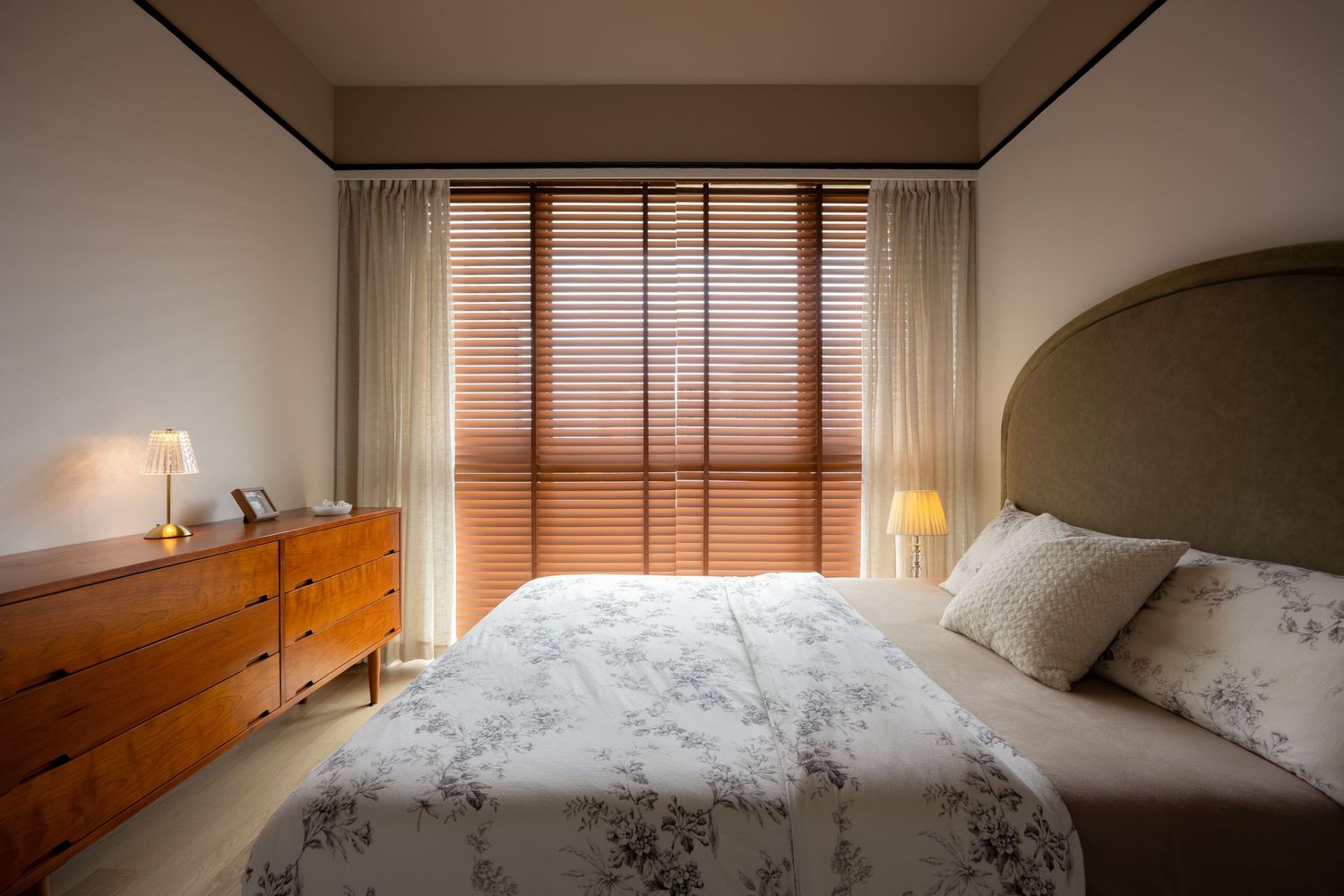 Cozy bedroom with floral bedding, wooden dresser, bedside lamps, and natural light filtering through wooden blinds.