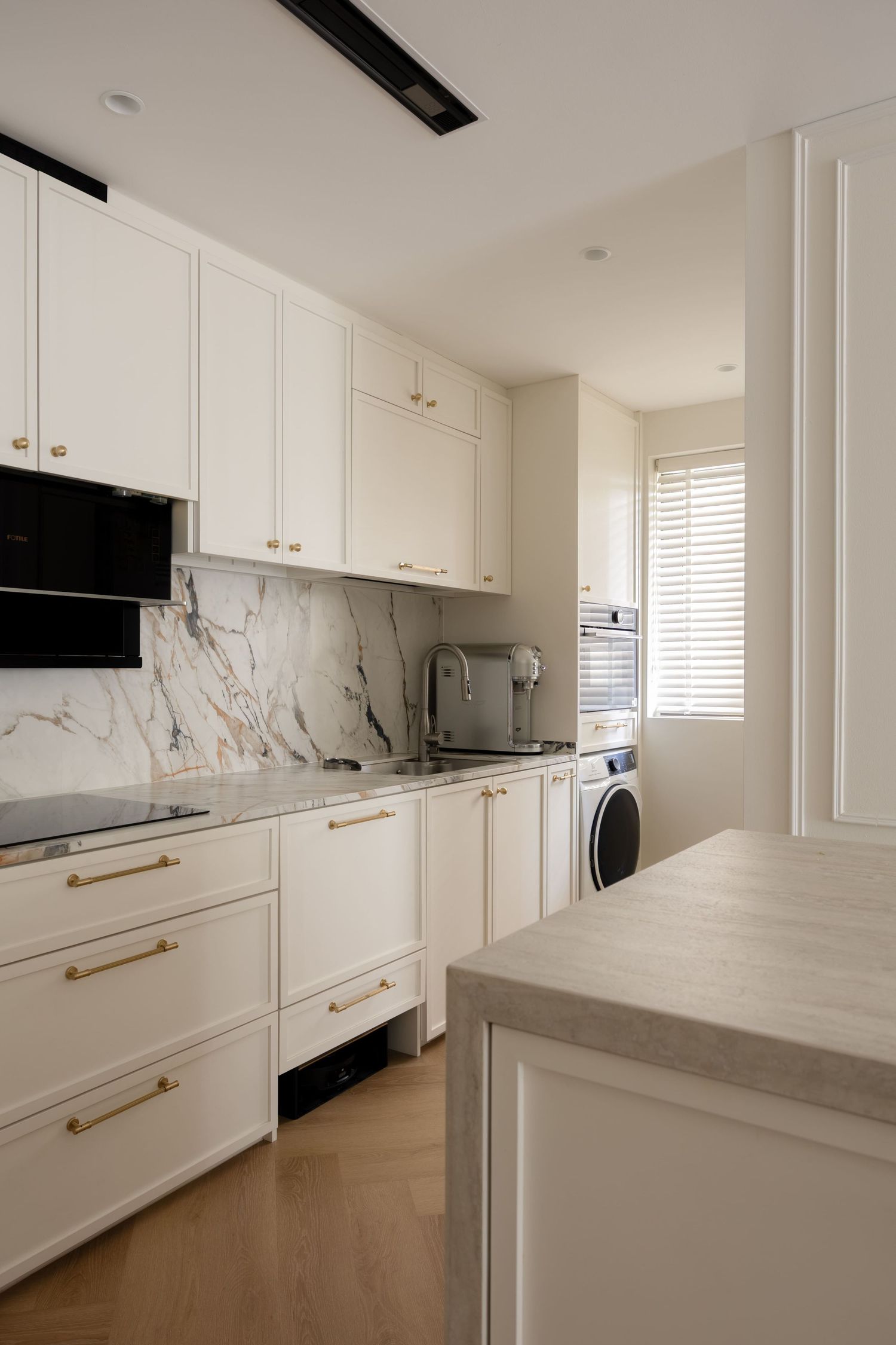 Modern white kitchen with marble backsplash, gold hardware, grey countertops, built-in appliances, and washer in utility area.