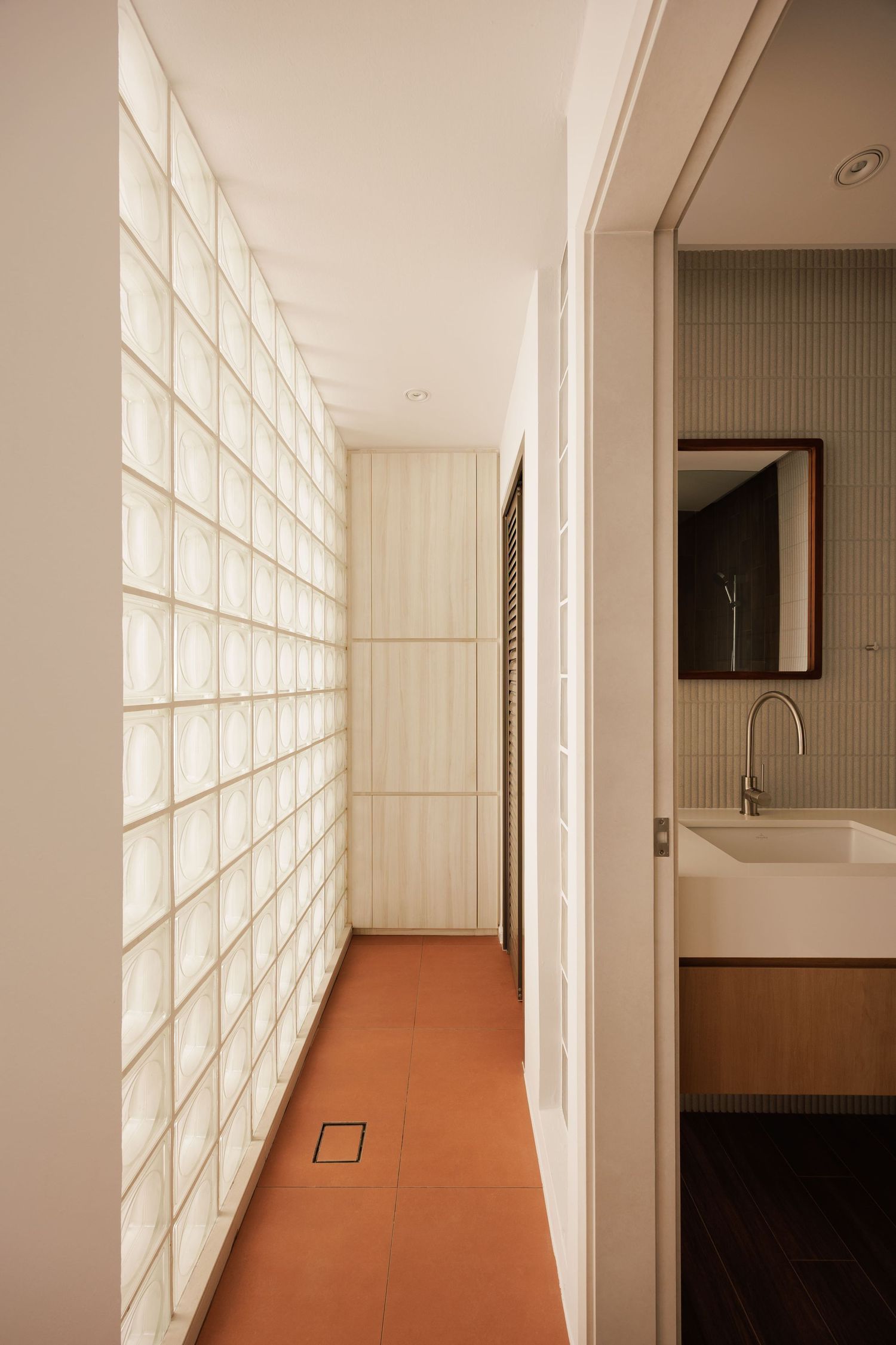 Modern interior hallway with glass block wall, terracotta floor tiles, and glimpse of minimalist bathroom with white sink.