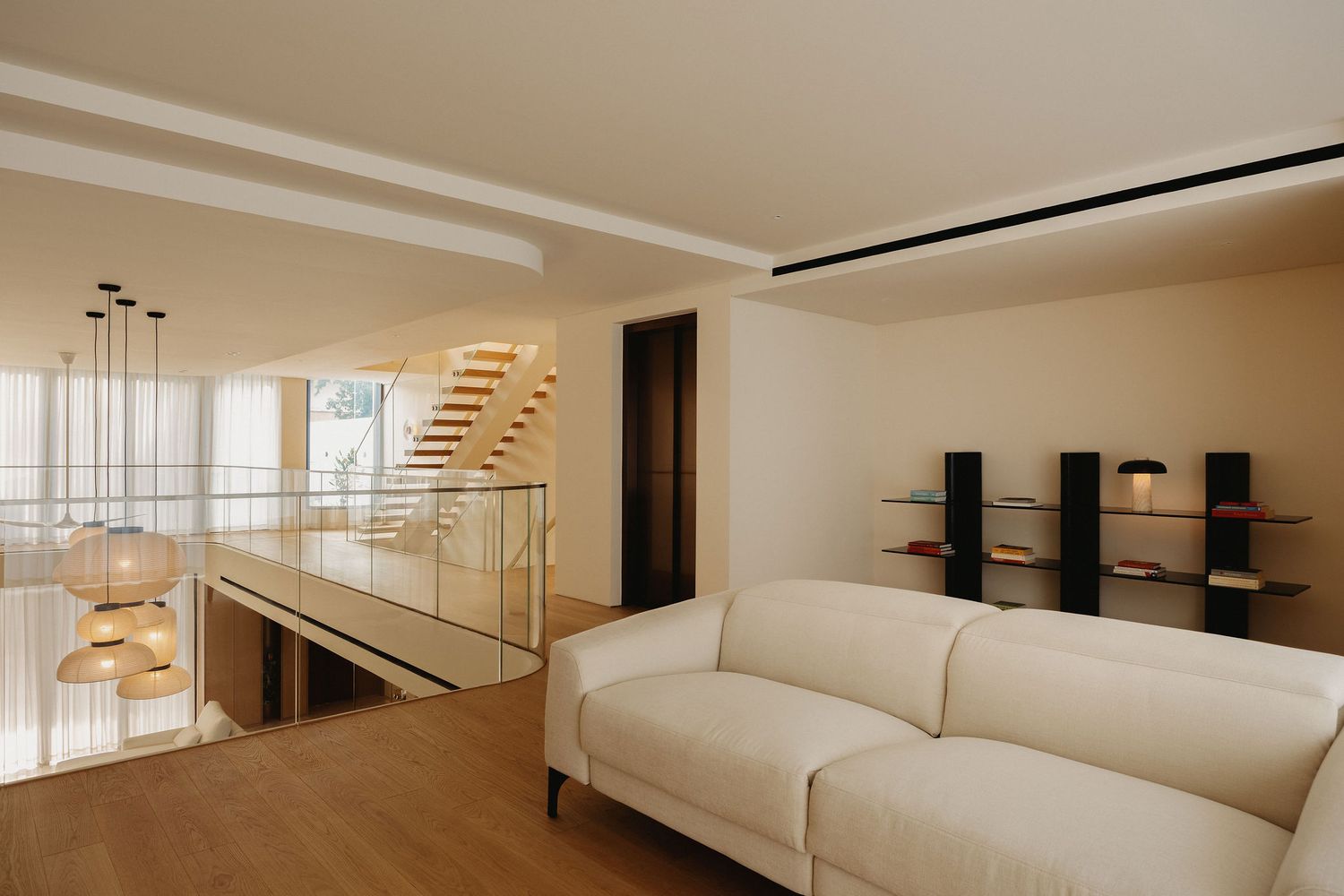 Modern minimalist living room with white sofa, floating staircase, glass railings, and sleek wall shelving on hardwood floors.