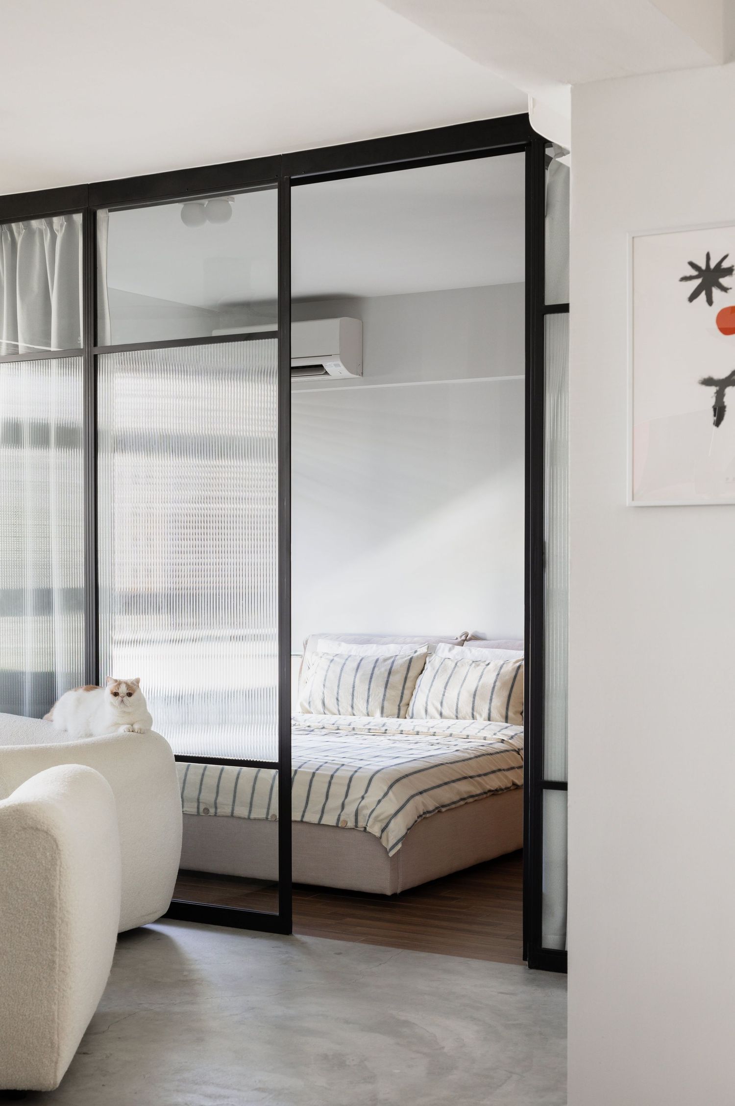 Modern bedroom with black-framed glass partition wall, white sofa, and striped bedding visible through sliding doors.