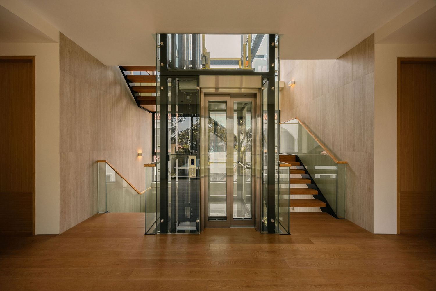 Modern glass elevator in a bright interior hallway with wooden staircases, glass railings and warm hardwood flooring.