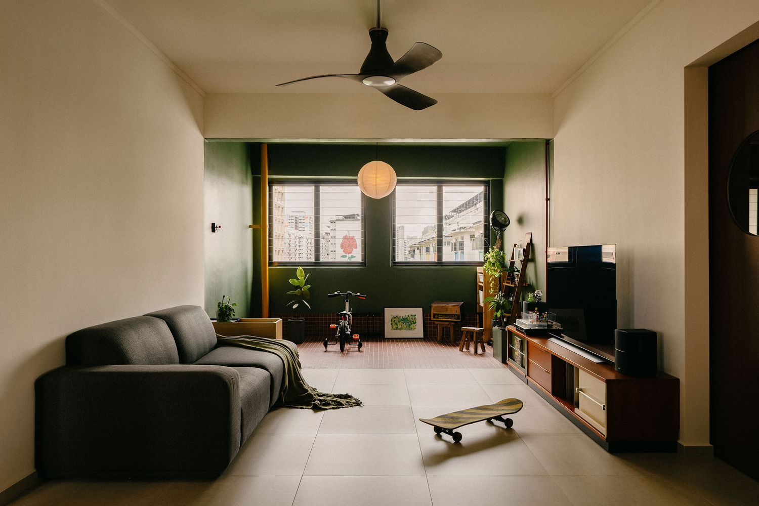 Modern living room with a gray sofa, ceiling fan, wooden TV console, green accent wall, pendant lamp, plants and a skateboard on the floor.