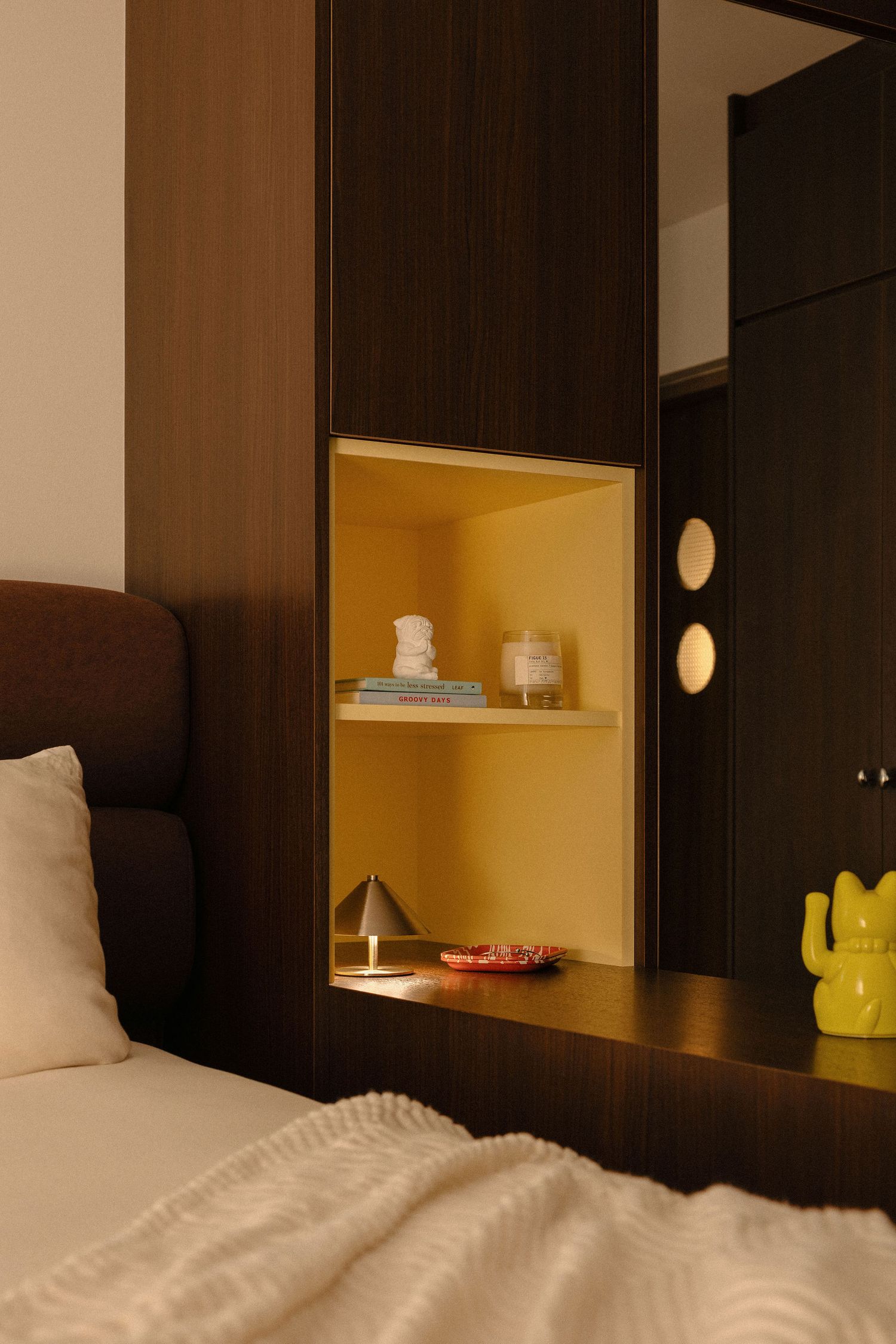 Cozy bedroom corner with dark wood cabinetry, a yellow built-in niche shelf, a small lamp, a lucky cat figurine, and soft cream bedding.