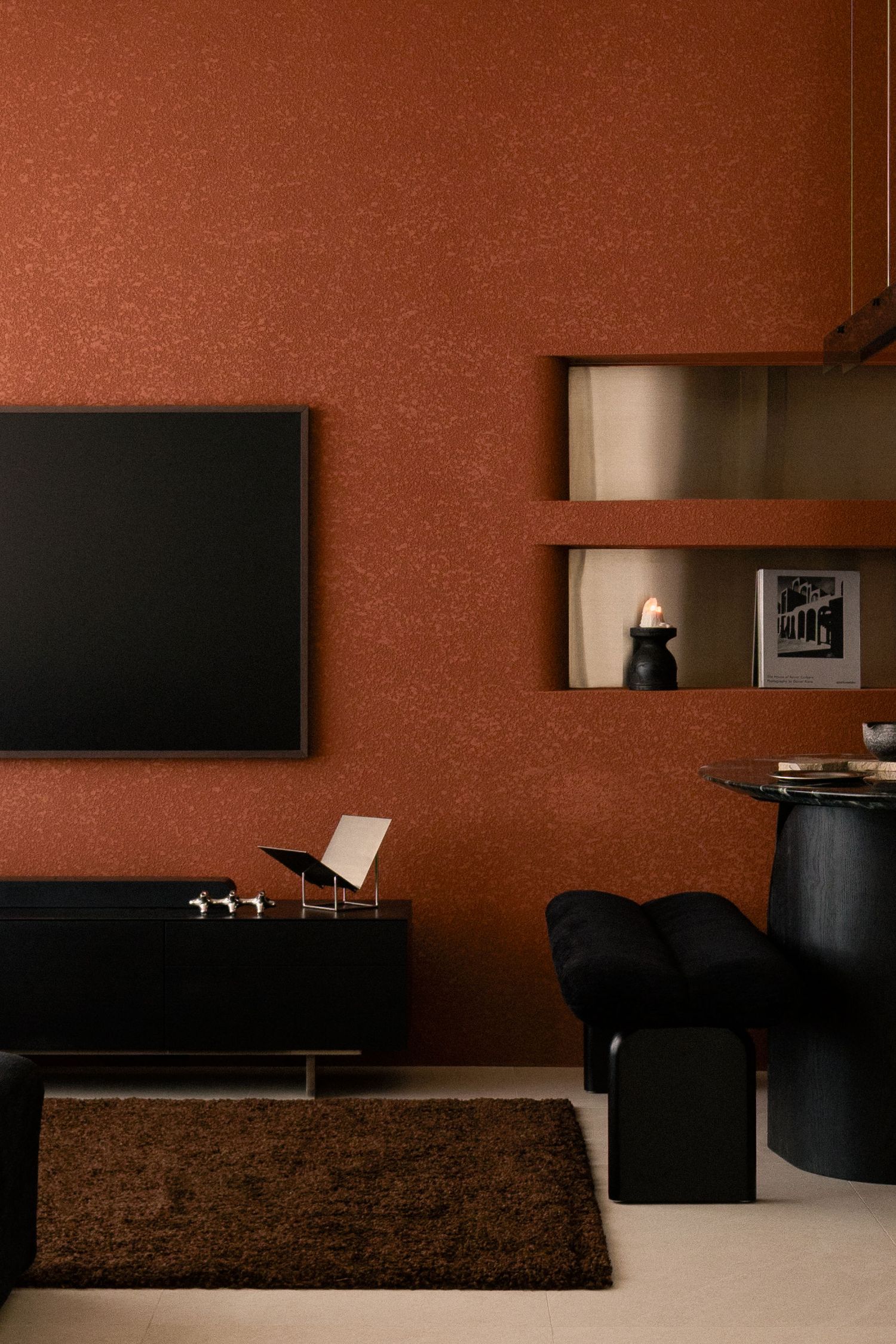 Modern living room with terracotta textured walls, wall-mounted TV, black sideboard, brass wall niches, shaggy brown rug and black stool.