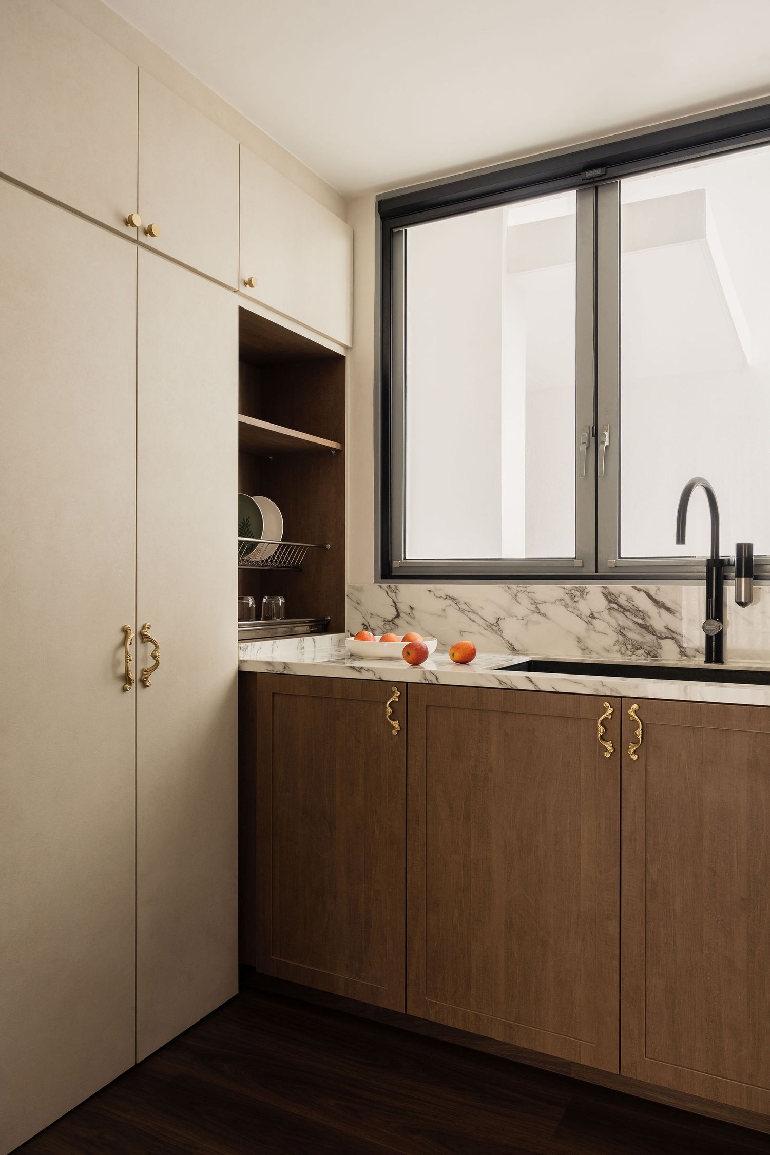 Modern kitchen with walnut cabinets, marble countertop, brass handles, black sink faucet, and cream upper cabinets near a large window.