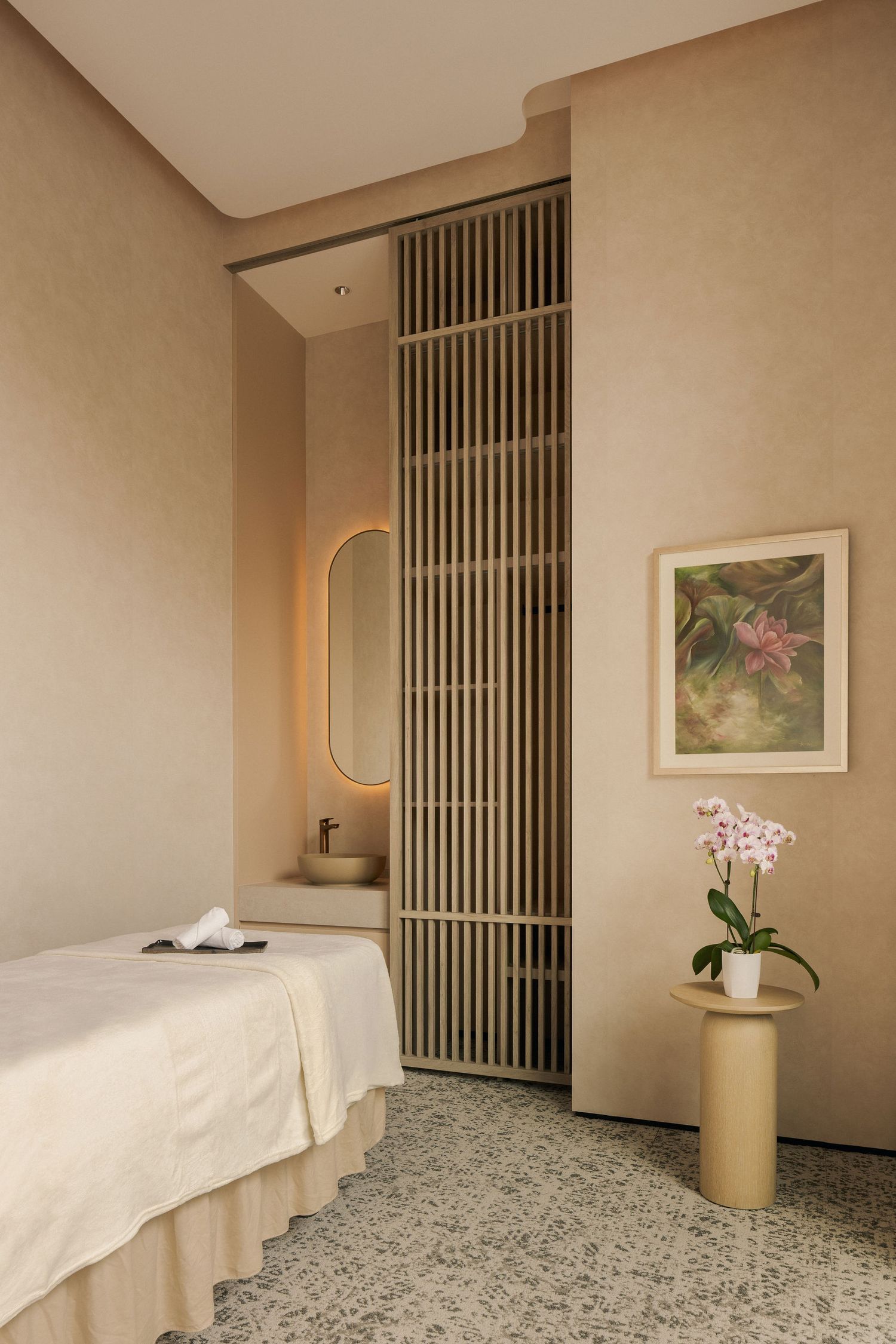 Serene spa treatment room with beige walls, massage table, wooden slat partition, oval mirror, and orchid on gold side table.
