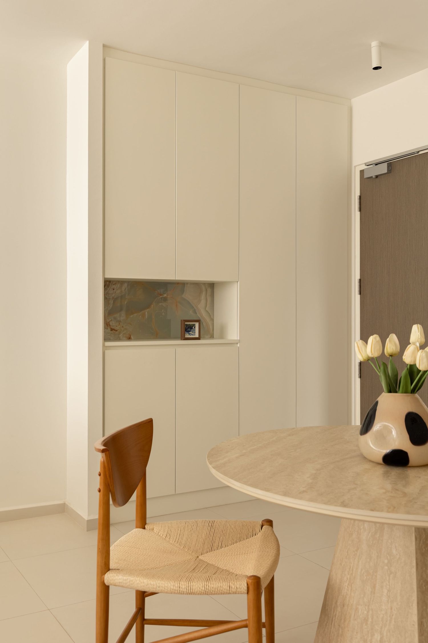 Modern minimalist dining area with wooden chairs, marble table, tulip vase, and built-in cream cabinetry with decorative niche.