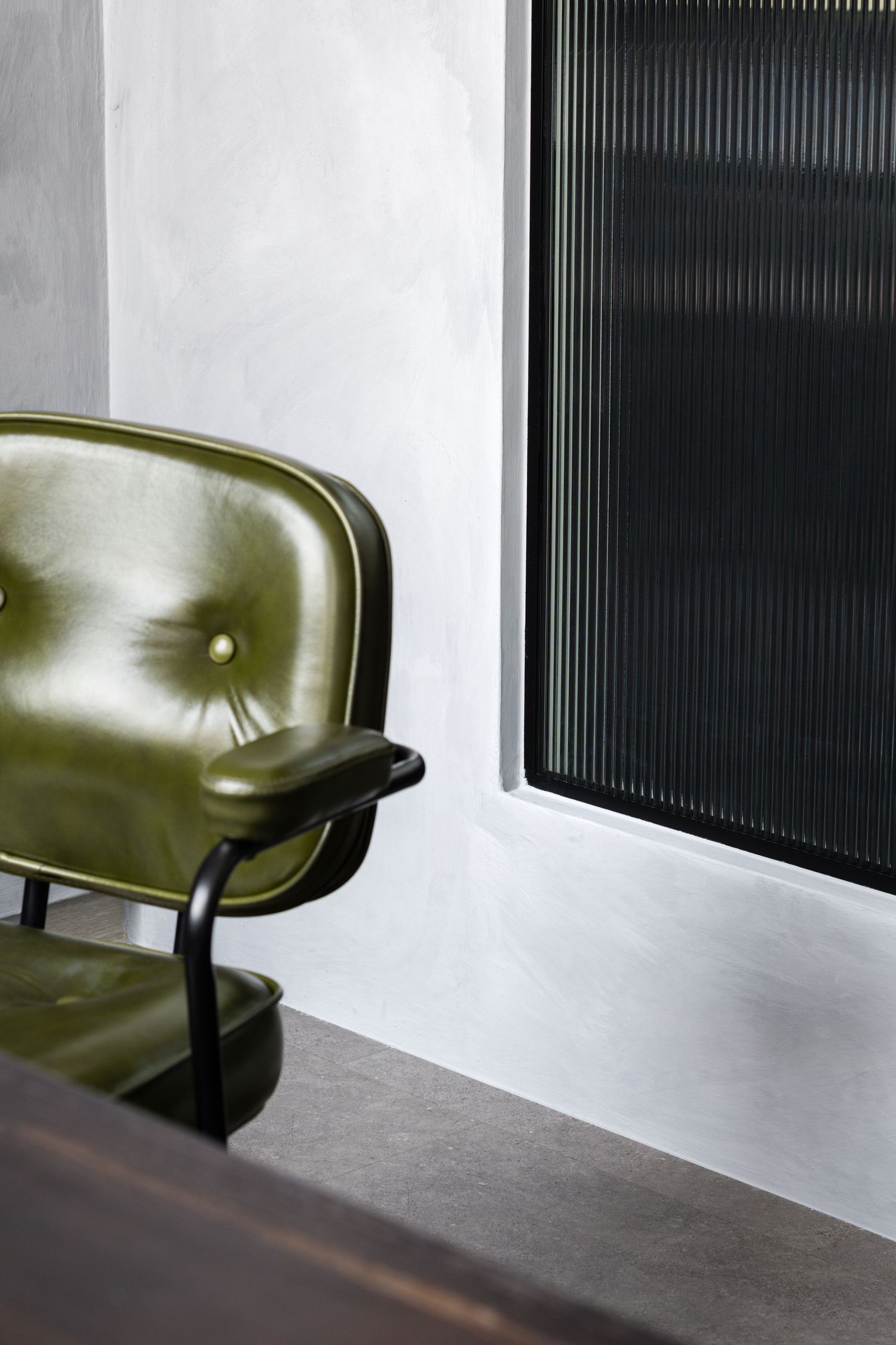 A vintage olive green leather chair sits against a white wall with a rectangular ribbed glass panel.