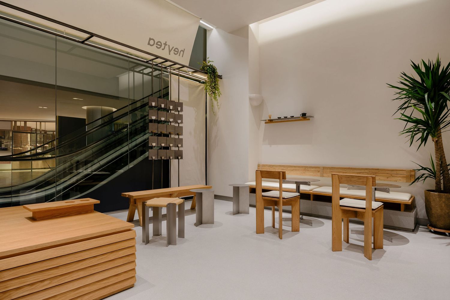 Minimalist cafe seating area with light wood tables, chairs and benches beside an escalator, white walls and a tall potted plant.