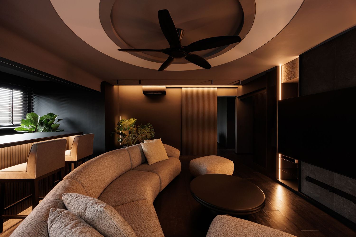Modern living room with curved beige sofa, ceiling fan, warm ambient lighting, and dark wood paneling.