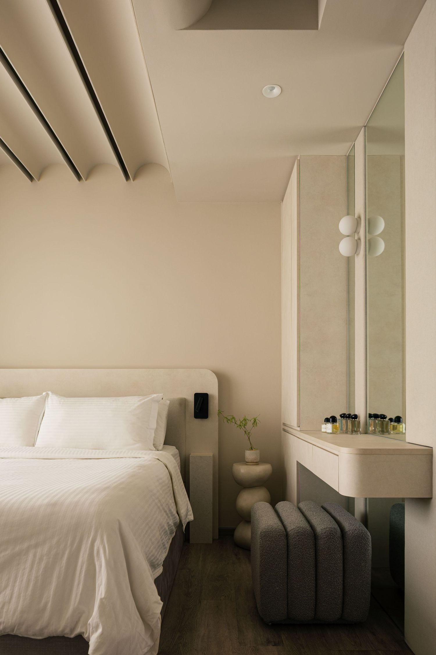 Minimalist hotel bedroom with white bedding, beige walls, vanity mirror with globe lights, and a gray tufted ottoman.
