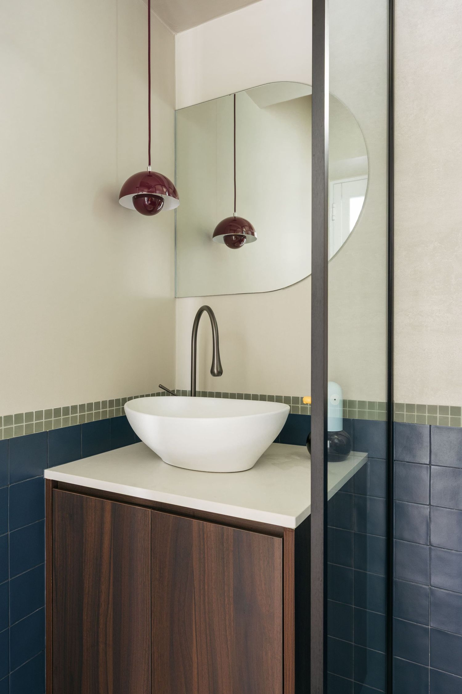 Modern bathroom vanity with white vessel sink, walnut cabinet, round mirror, and burgundy pendant lights against tiled walls.