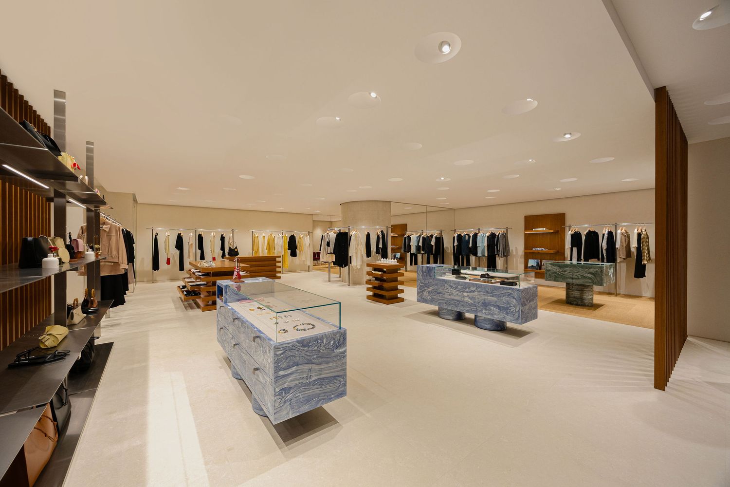 Spacious luxury retail store interior with marble display tables, clothing racks, and warm beige tones.