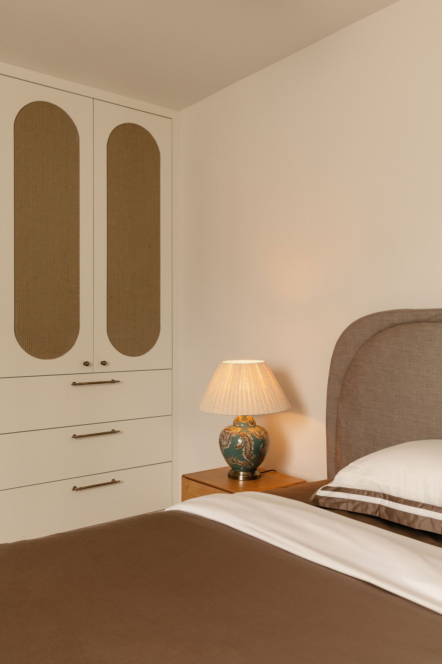 Elegant bedroom with cream wardrobe, green ceramic table lamp on wooden nightstand, and upholstered headboard.