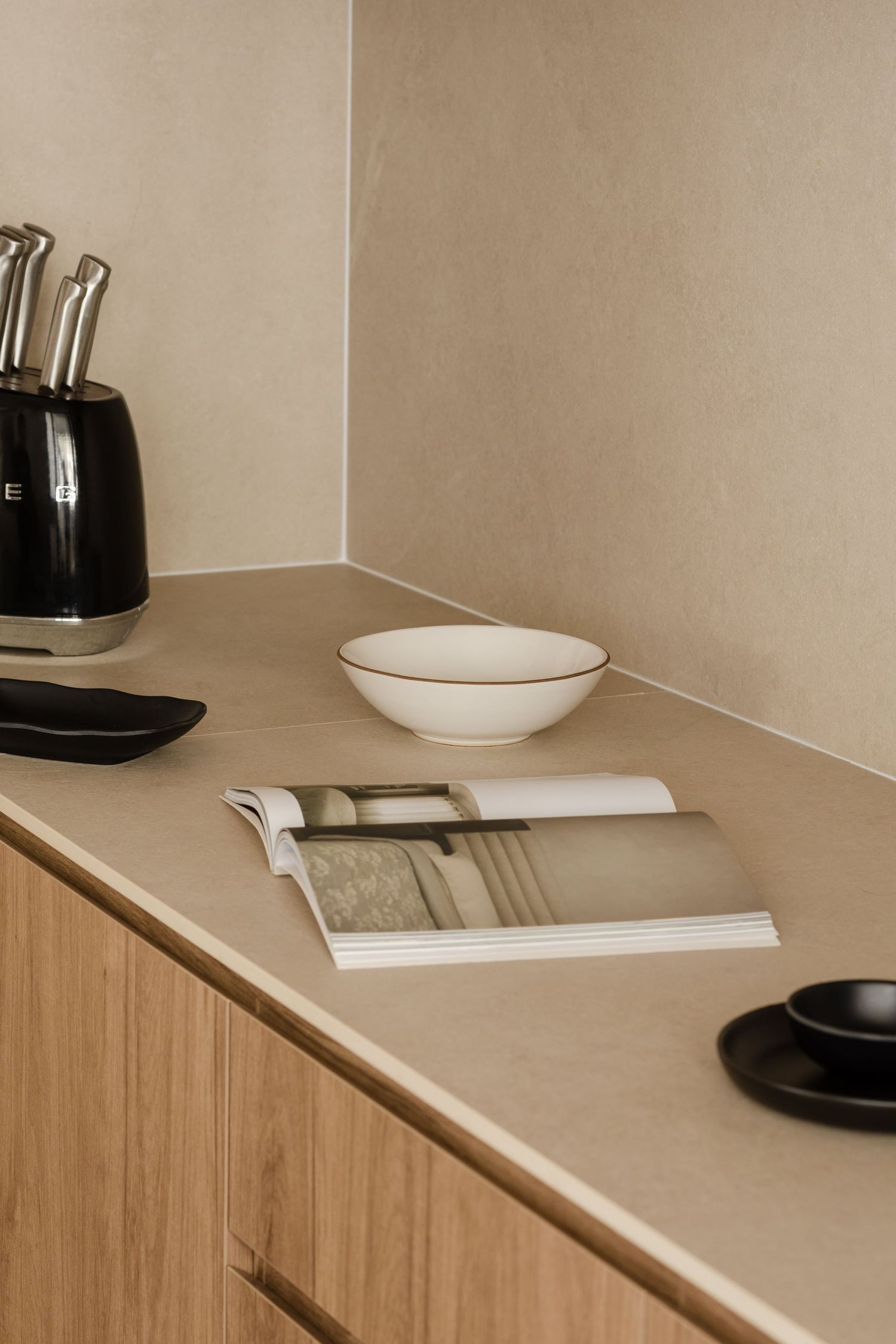 Minimalist kitchen counter with wooden cabinets, beige countertop, knife set, open book, and simple white bowl.