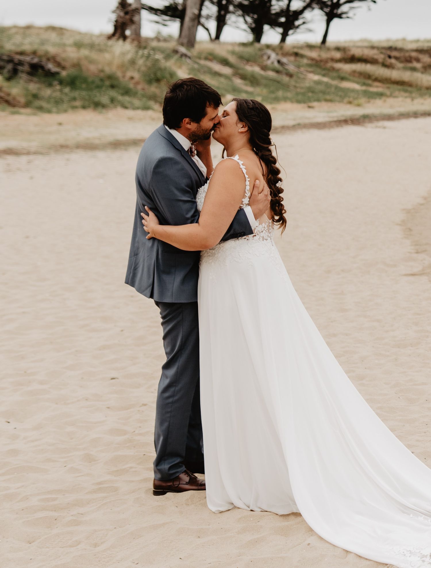 A couple shares a romantic kiss on a sandy beach, the bride in a flowing white gown and groom in a gray suit.
