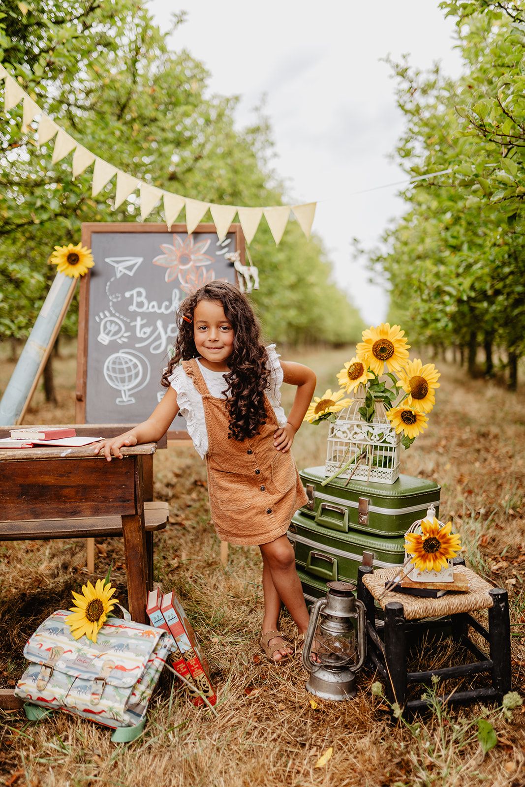 Girl in brown overalls poses by chalkboard with sunflowers and vintage suitcases in an orchard for back to school.