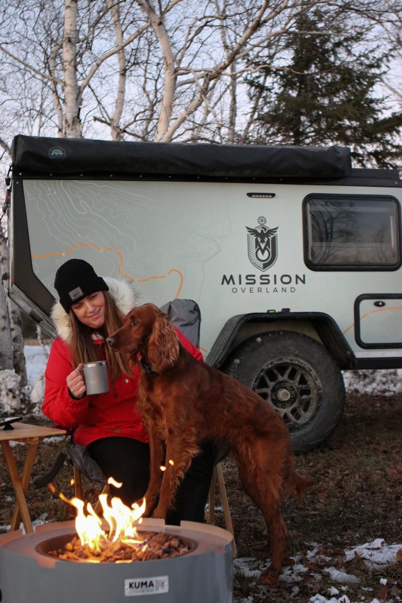 Person enjoying coffee with their brown dog by a campfire next to a Mission camper trailer.