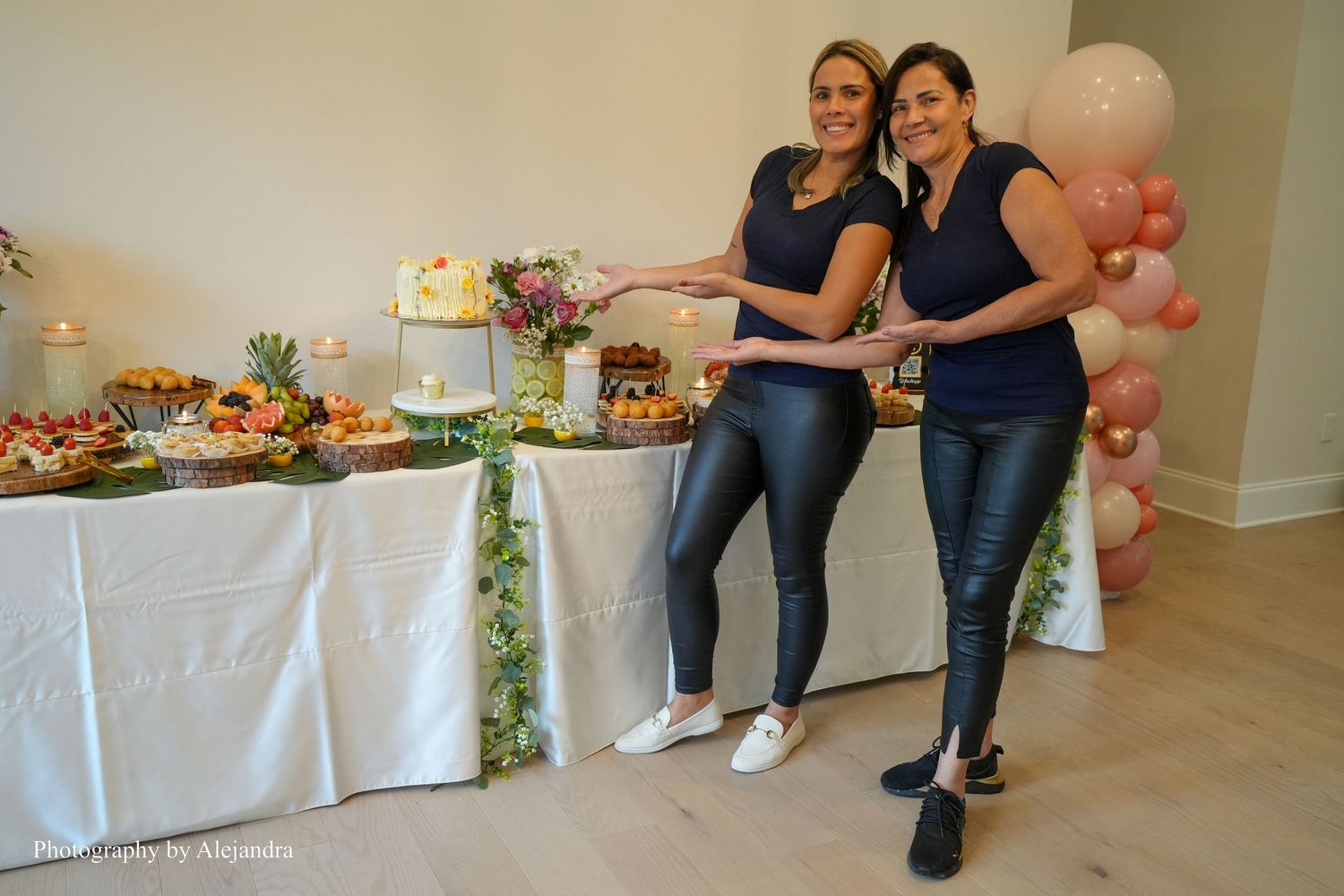 Two women in matching black outfits smiling beside elegant dessert table with pink balloon arch.