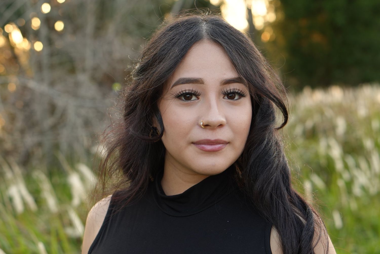 Young Latina woman with long dark wavy hair poses outdoors with golden grass background at sunset.