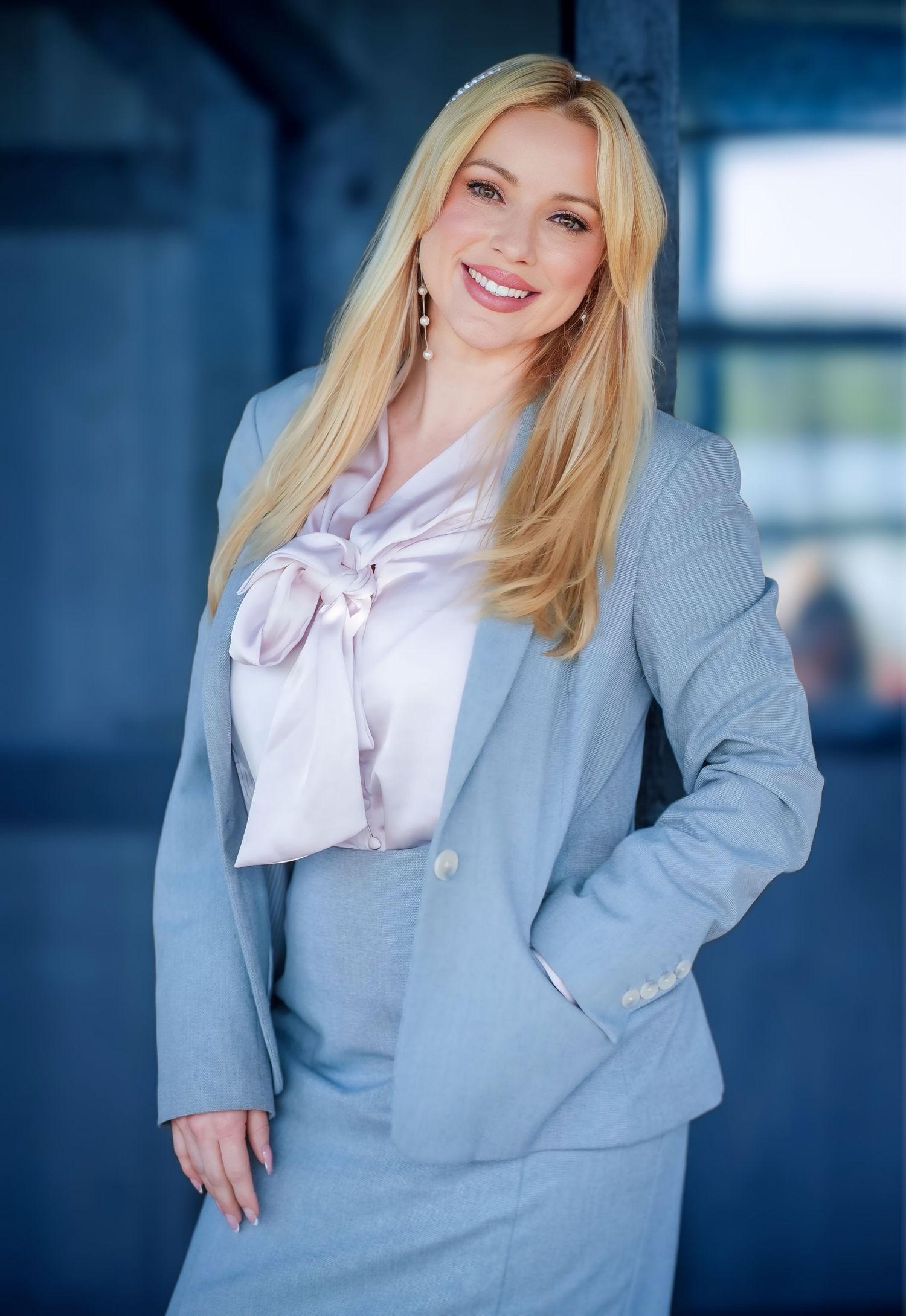 Professional blonde woman smiling in a light gray business suit with white blouse, posing confidently indoors.