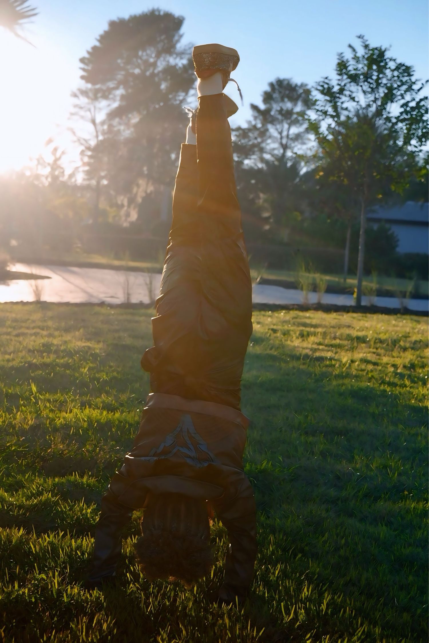Person doing a headstand on green grass lawn in bright sunlight with trees in background.
