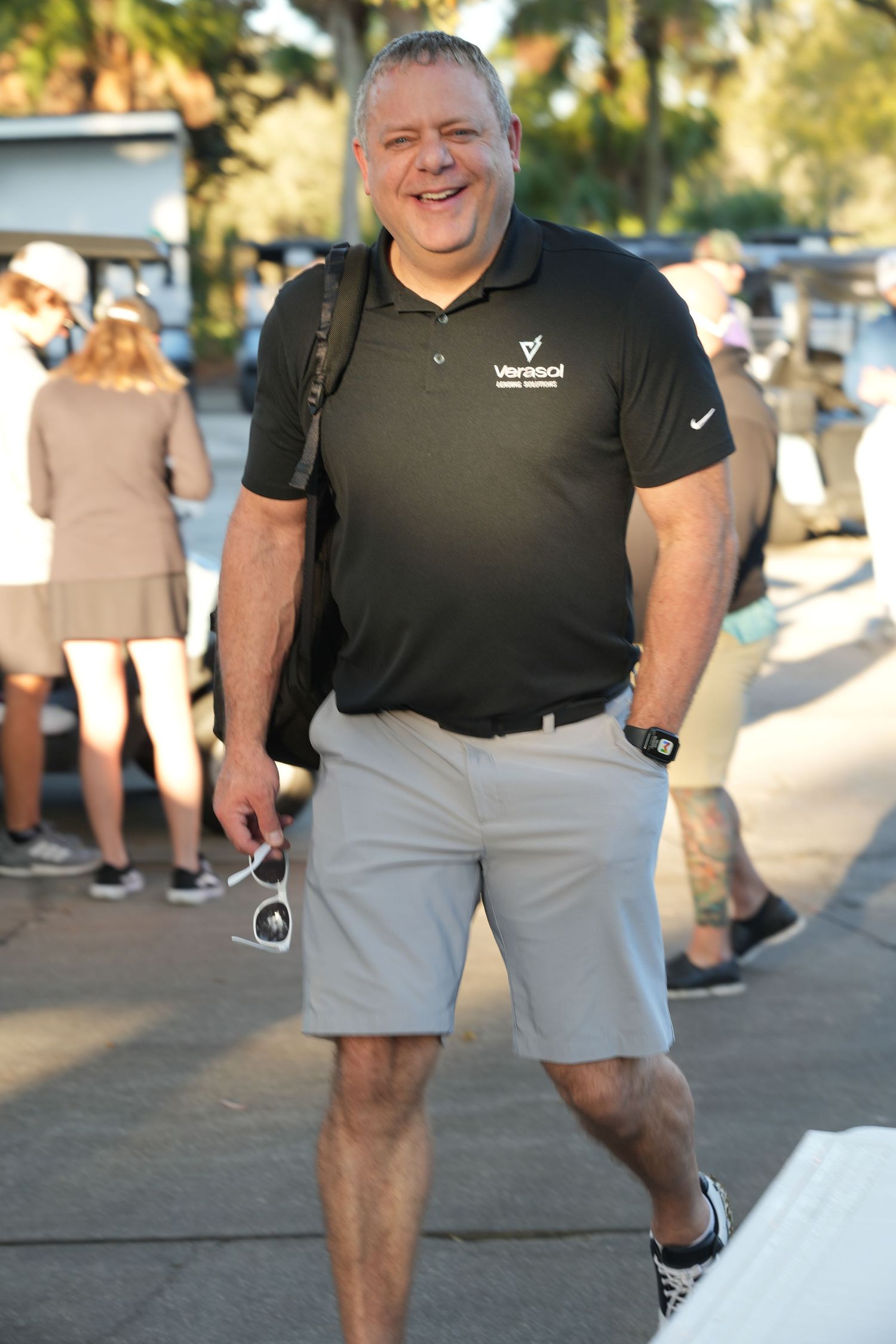 Smiling man in black Varsol polo shirt and khaki shorts standing outdoors at casual event.