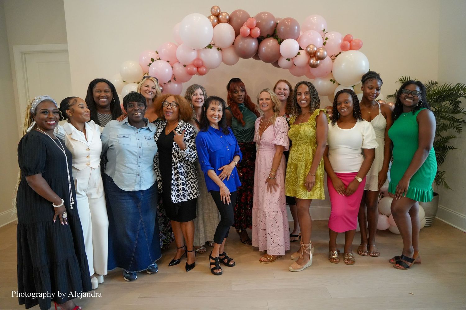 Group of diverse women posing together indoors under pink balloon arch at celebration event.