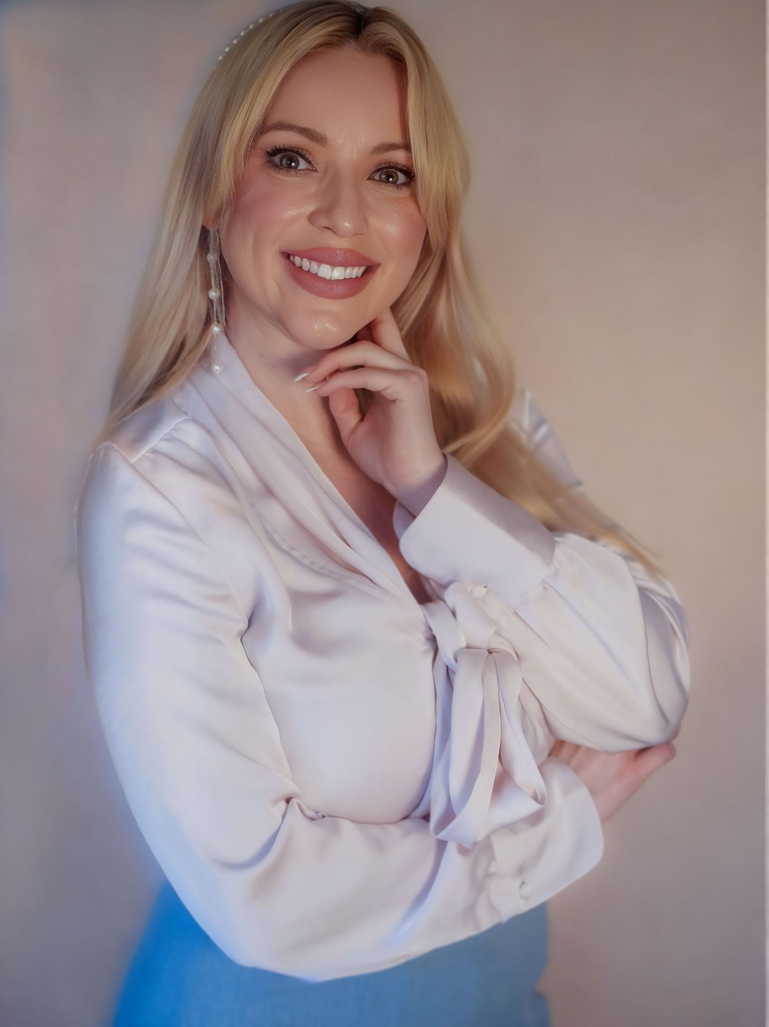 Blonde woman in white satin blouse smiles warmly with hand on chin against neutral backdrop.