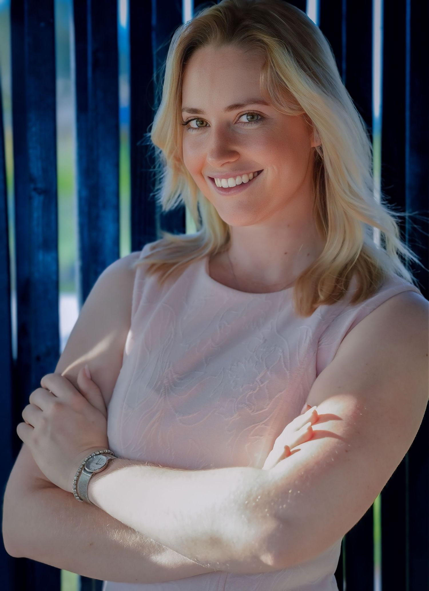 Smiling blonde woman in pink top with arms crossed posing in front of blue striped background.