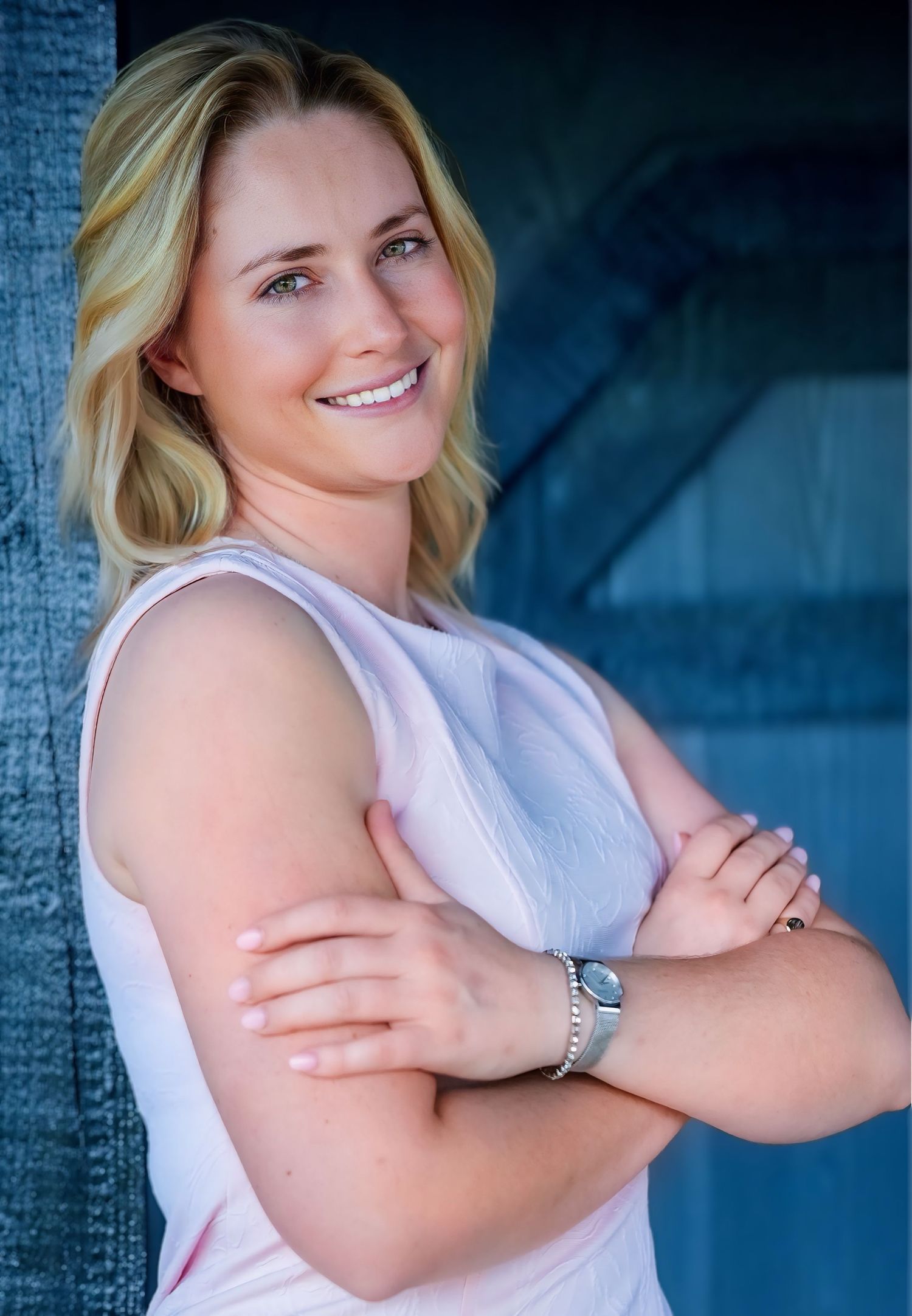 Smiling blonde woman in white sleeveless top with arms crossed poses against blue background.