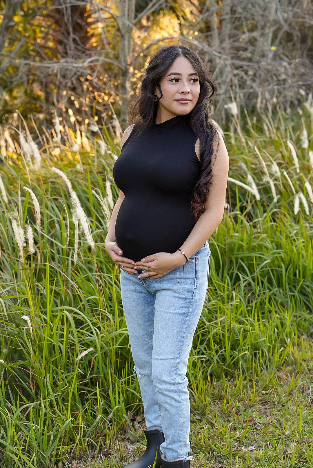 Pregnant woman in black sleeveless top and jeans holds baby bump outdoors among tall green grass.