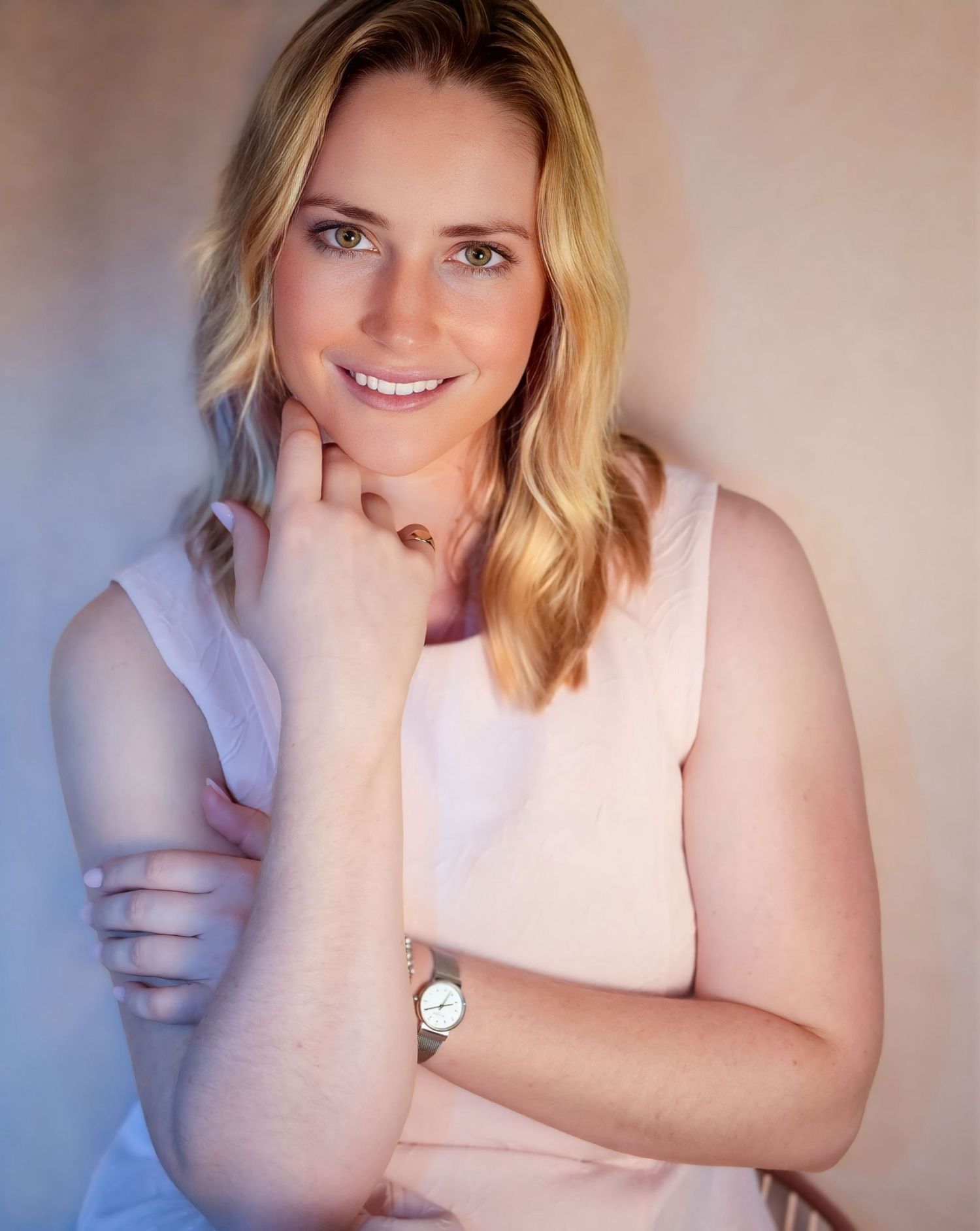 Smiling blonde woman in pink sleeveless top poses with arms crossed in warm lighting.
