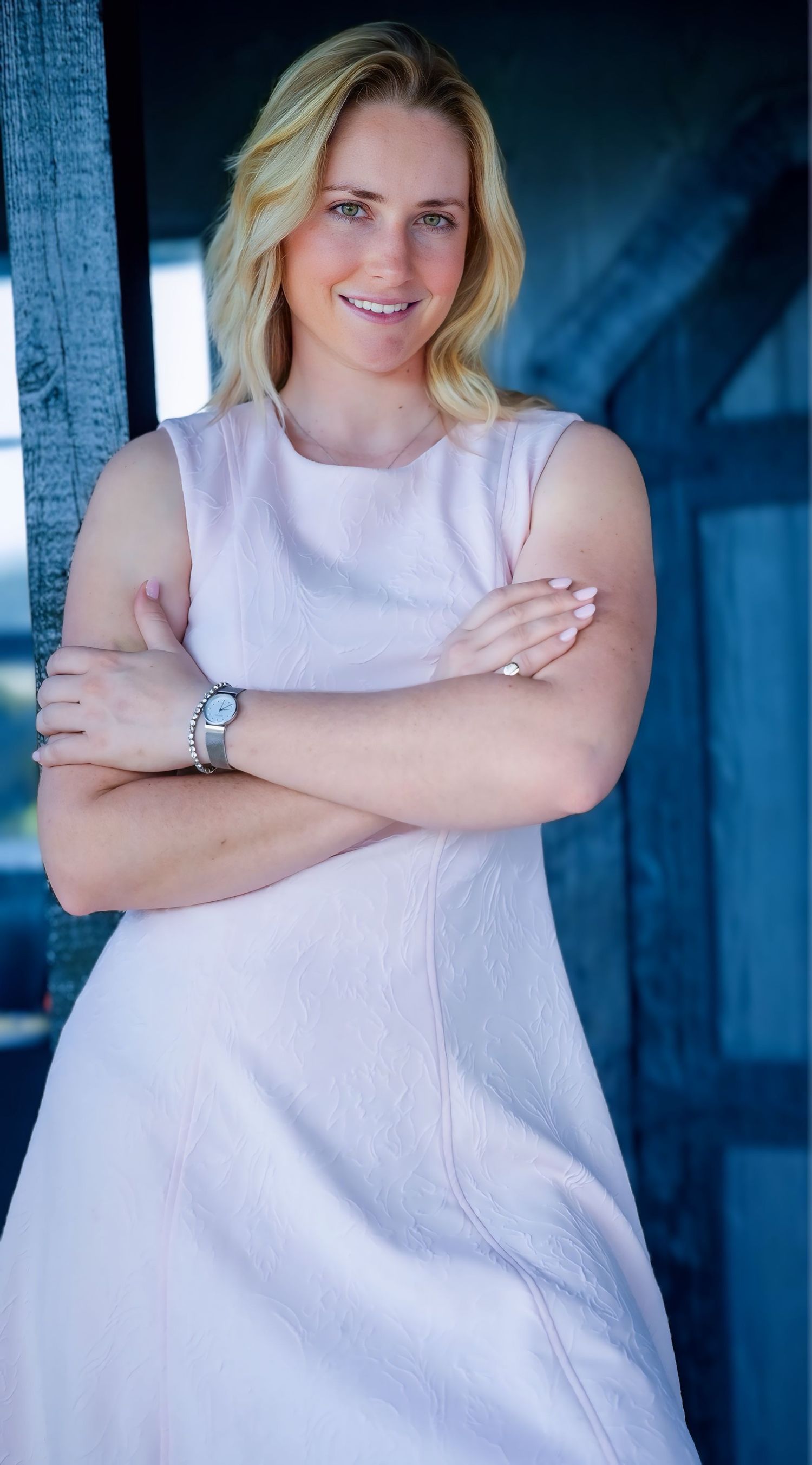 Smiling blonde woman in pink sleeveless dress stands with arms crossed in elegant outdoor setting.